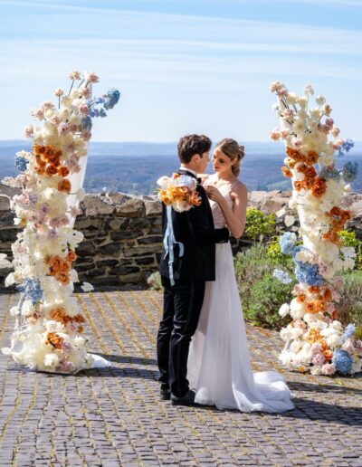 Brautpaar steht am Meer unter einem Blumenbogen, beide halten sich nah und lächeln