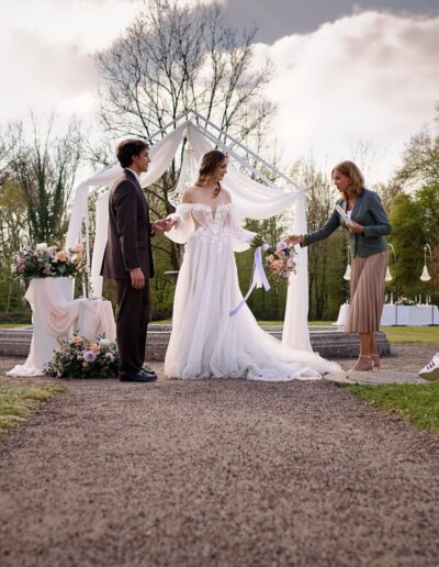 Brautpaar steht vor Traurednerin im Freien während einer Trauung, Stühle und Gäste im Hintergrund